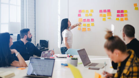 Menschen in einem Meetingraum. Eine Person steht vor einer Wand mit Haftnotizen.