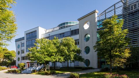 Hochschulgebäude von der Straße aus gezeigt. Davor befinden sich ein paar Bäume und eine Straße.